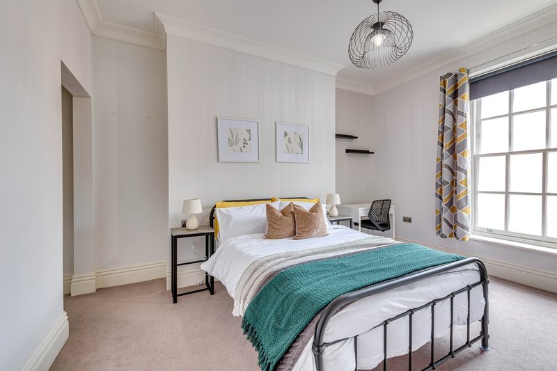 Modern bedroom with double bed, metal frame, turquoise throw, white bedding, wooden nightstand, window with patterned curtains, pendant light, and framed wall art