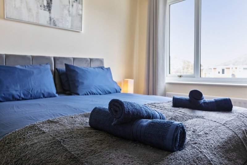 Modern bedroom with blue bedding, rolled towels on bed, and large window with city view