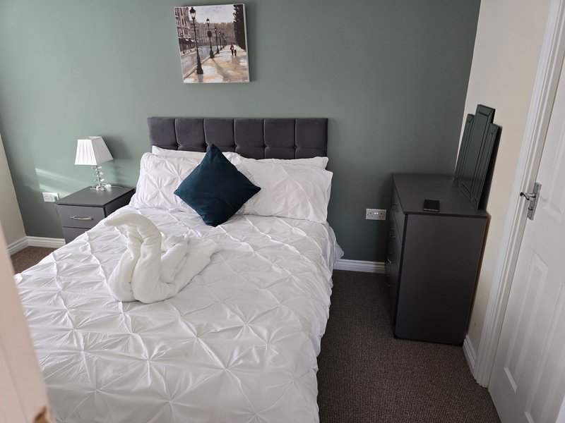 Bedroom with double bed, grey headboard, white bedding, dark cushion, bedside lamp and wall art