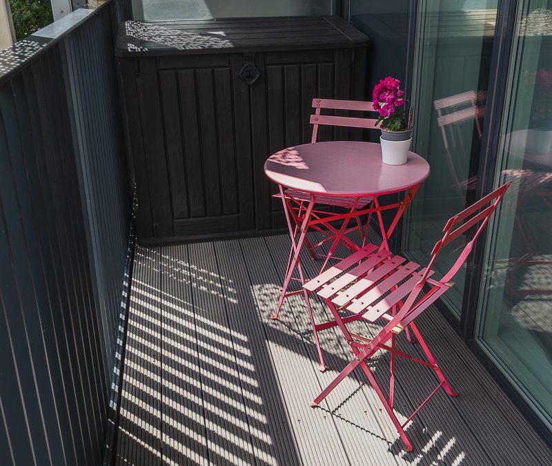 Small outdoor patio with pink round table, two pink chairs, potted magenta flower, black gate and decking. Space to store tools on the balcony.