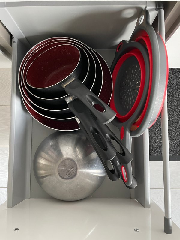 Kitchen drawer containing stacked cookware including pans, red colanders, and a metal pot