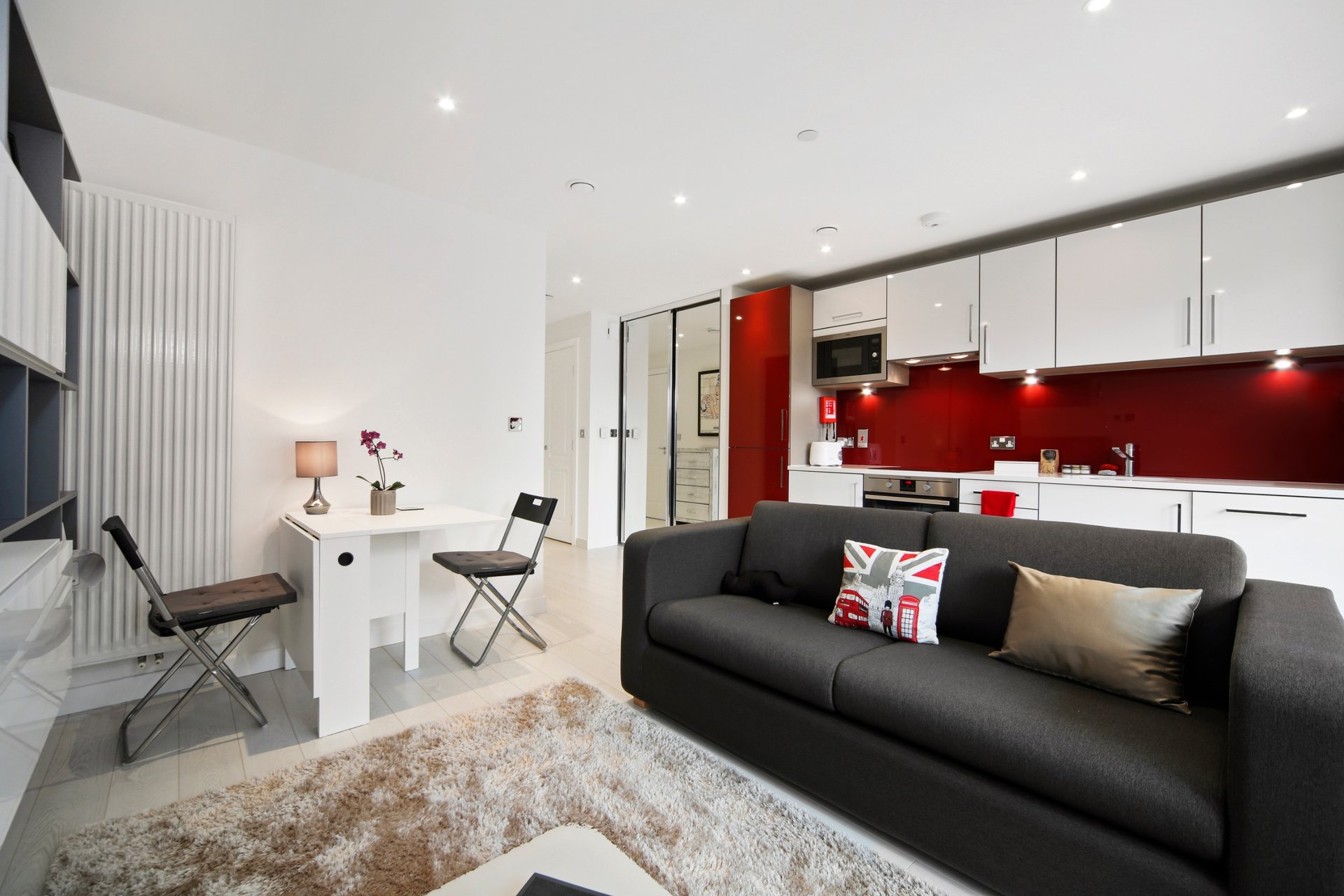 Open-plan living room with black sofa, white desk, red kitchen backsplash and recessed lighting