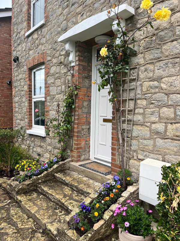Stone cottage entrance with white door, flowering hanging basket, and colorful garden beds
