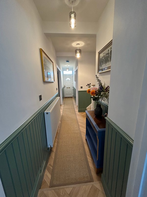 Long hallway with green wainscoting, pendant lights, framed artwork, and potted flowers on console table