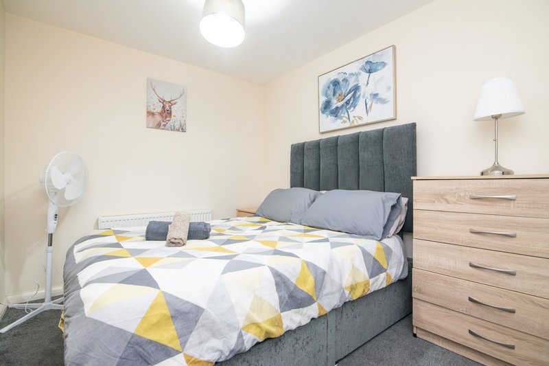 Bedroom with double bed, grey upholstered headboard, geometric patterned bedding, wooden dresser and wall art
