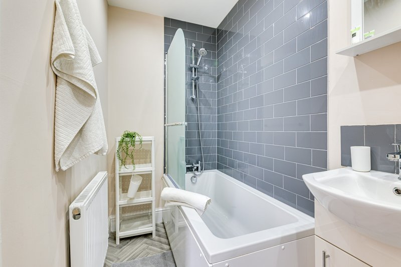 Modern bathroom with white bathtub, grey tile surround, white sink and wooden shelving unit