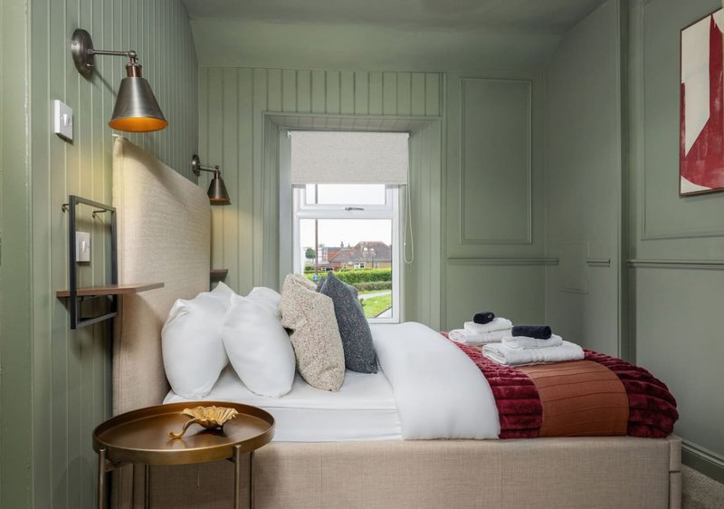 Bedroom with single bed, white pillows, red blanket, wall sconces and green panelled walls