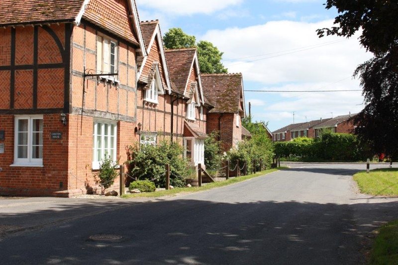Row of brick and timber-framed cottages with driveways, green gardens and trees lining quiet street