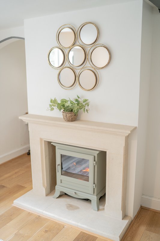 Living room fireplace with cream mantel, decorative mirror arrangement, potted plant, and wood stove insert