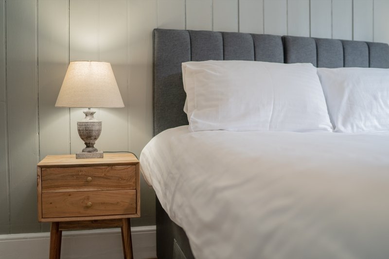 Modern bedroom with grey upholstered bed headboard, white bedding, wooden bedside table and table lamp