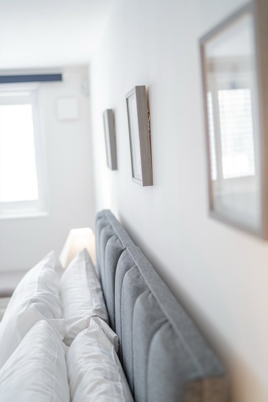 Bright bedroom with white and grey bedding, wall-mounted heating unit, and windows