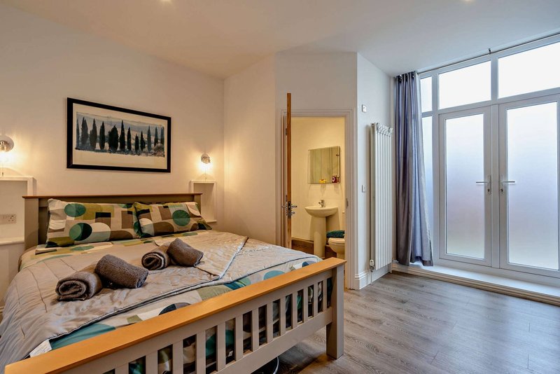 Bright bedroom with wooden bed, patterned bedding, large windows, and ensuite bathroom visible