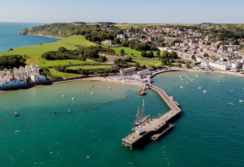 Aerial view of coastal town with beach, harbor pier, boats and green cliffs