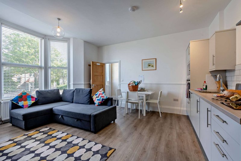 Open-plan living area with blue corner sofa, dining table, kitchen, and patterned rug