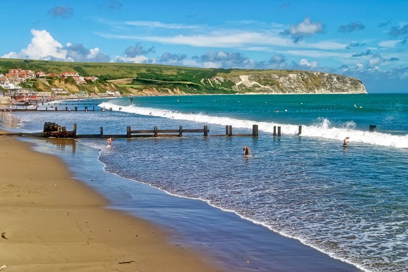 Coastal beach scene with golden sand, blue sea, wooden pier, and green cliffs under clear sky