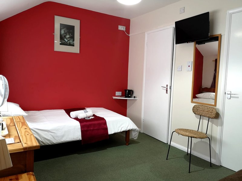 Bedroom with red accent wall, single bed with white linens, mirror, and wicker chair