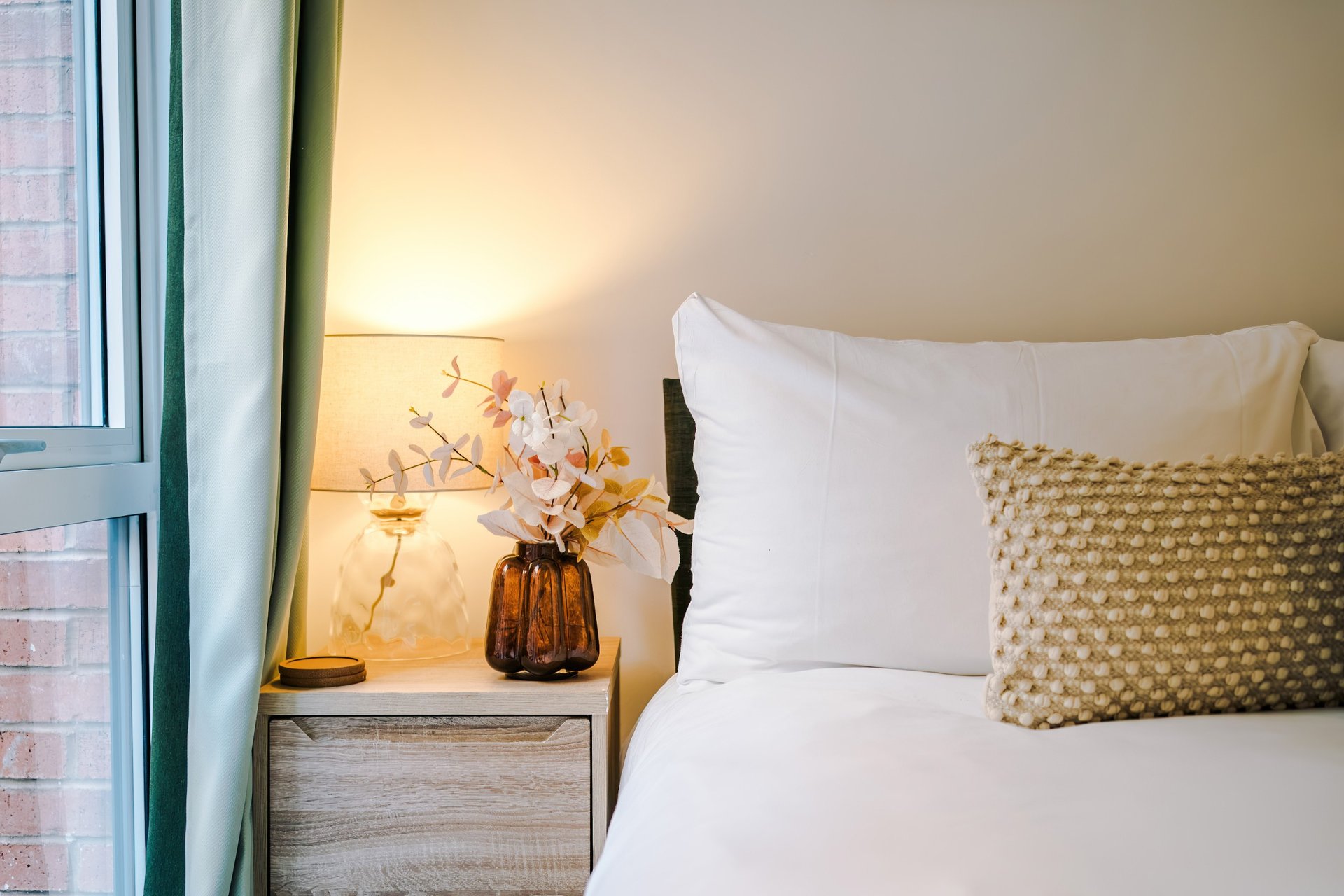 Bedroom with white pillows, patterned cushion, bedside lamp and dried flowers in vase