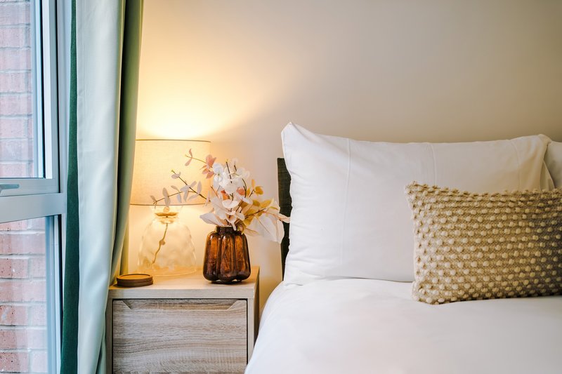 Bedroom with white pillows, patterned cushion, bedside lamp and dried flowers in vase
