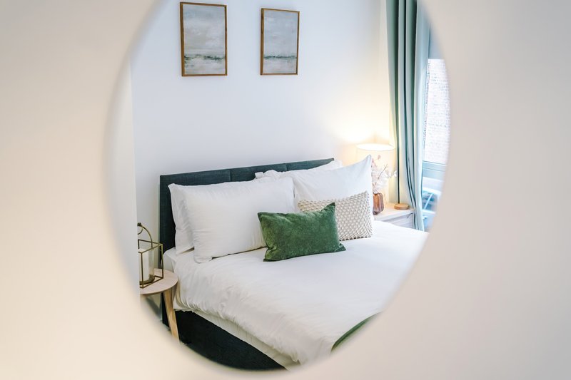 Bright bedroom with double bed, green and white pillows, framed wall art, and round mirror