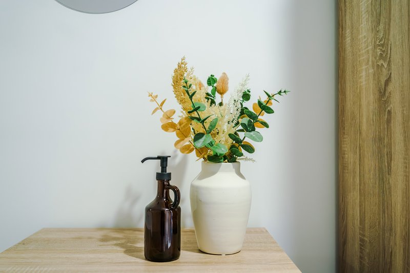 Wooden desk with white ceramic vase containing dried flowers and dark glass soap dispenser