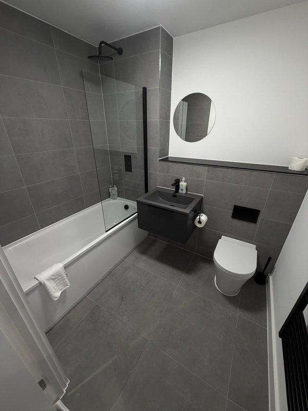 Modern bathroom with grey tile walls, white bathtub, black vanity sink, toilet, and round mirror