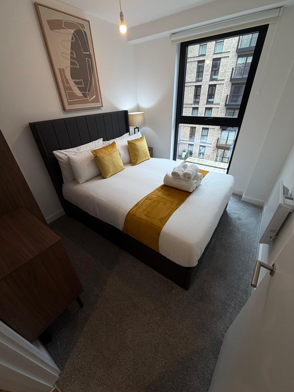 Modern bedroom with double bed, yellow accent cushions, framed wall art and large window overlooking urban street