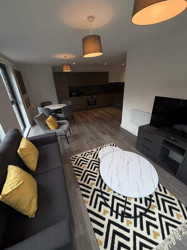 Open-plan living space with dark sofa, yellow cushions, white marble table, and kitchen area