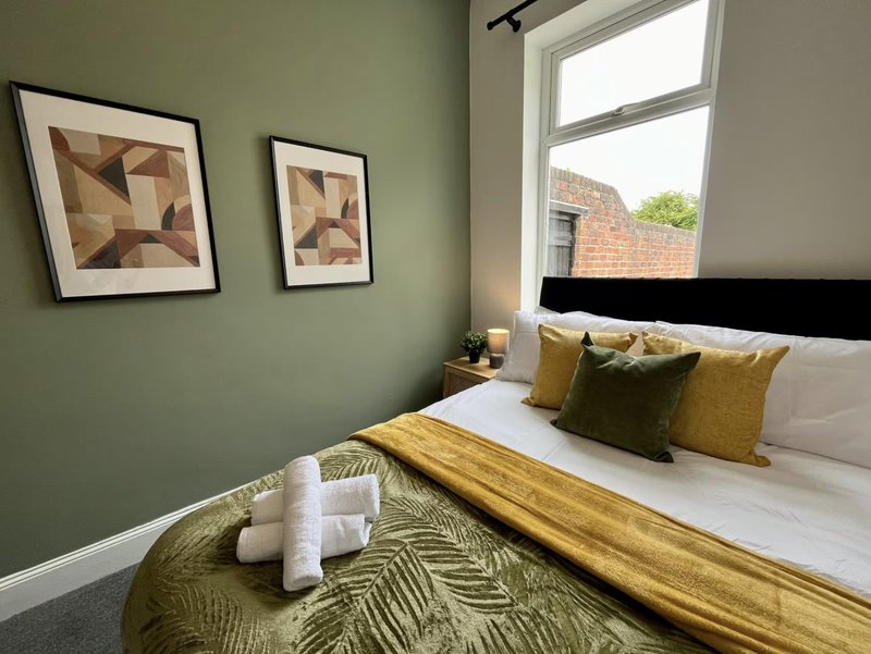 Bedroom with sage green walls, double bed, gold bedding, framed abstract art, and bright window.