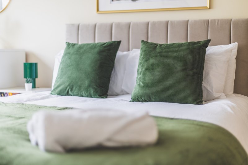 Bedroom with grey upholstered headboard, green pillows, white bedding and bedside lamp