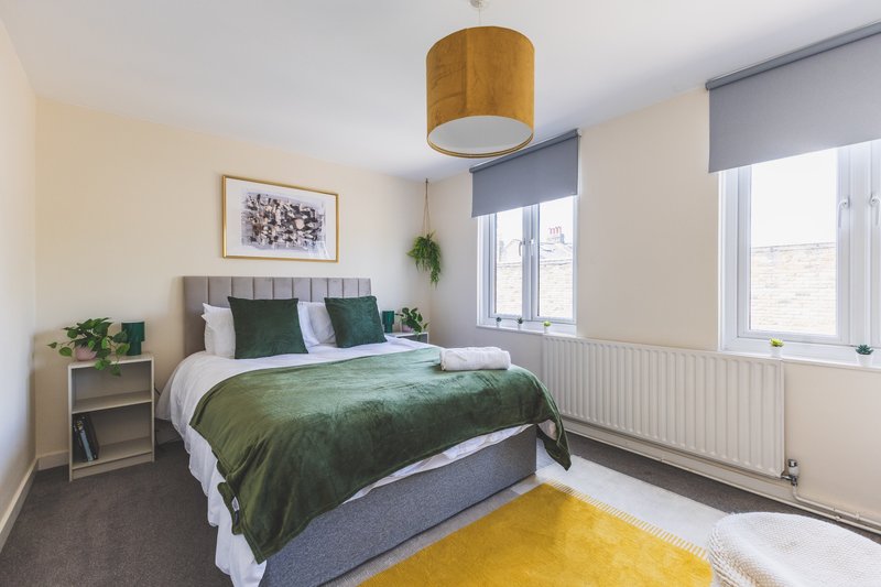 Bright modern bedroom with double bed, green bedding, wooden ceiling lamp, and yellow rug