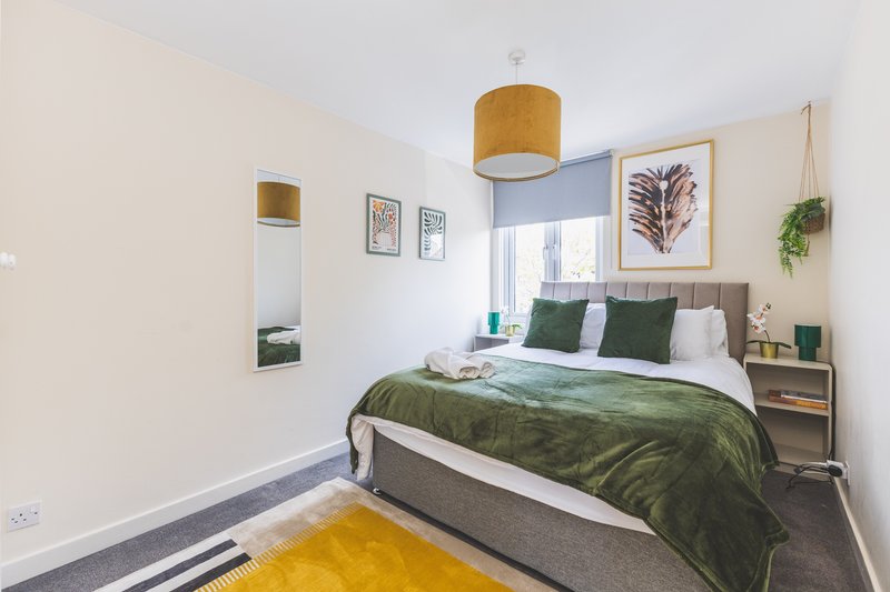 Modern bedroom with double bed, green bedding, wooden pendant lamp, framed wall art and yellow rug
