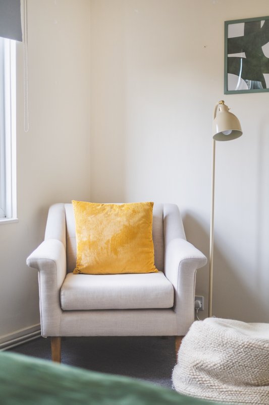Bright bedroom corner with cream armchair, yellow cushion, floor lamp and window