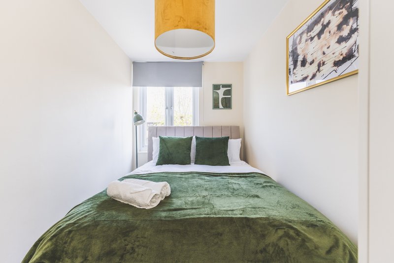 Narrow bedroom with double bed, green bedding, wooden ceiling lamp and framed wall art