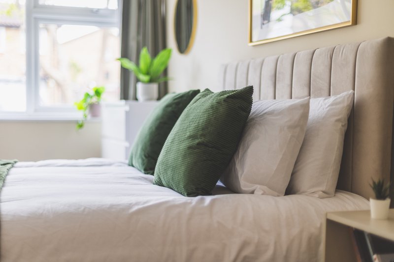 Modern bedroom with made bed, green and cream pillows, upholstered headboard, and window with plants