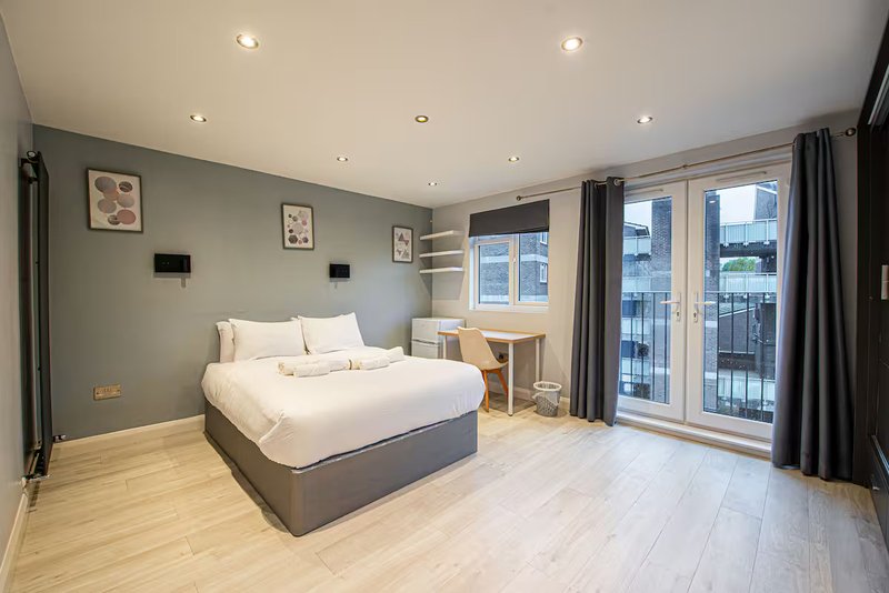 Modern bedroom with double bed, wooden floor, desk, and balcony doors overlooking city