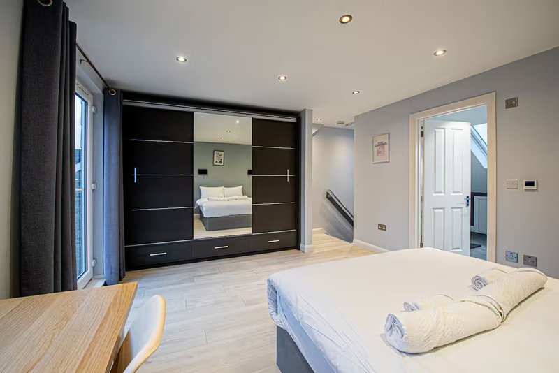 Modern bedroom with bed, wooden desk, large black wardrobe with mirror doors, and en-suite bathroom visible through open door