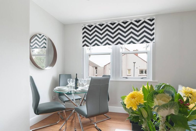 Modern dining area with glass table, grey chair, round mirror and chevron patterned blind