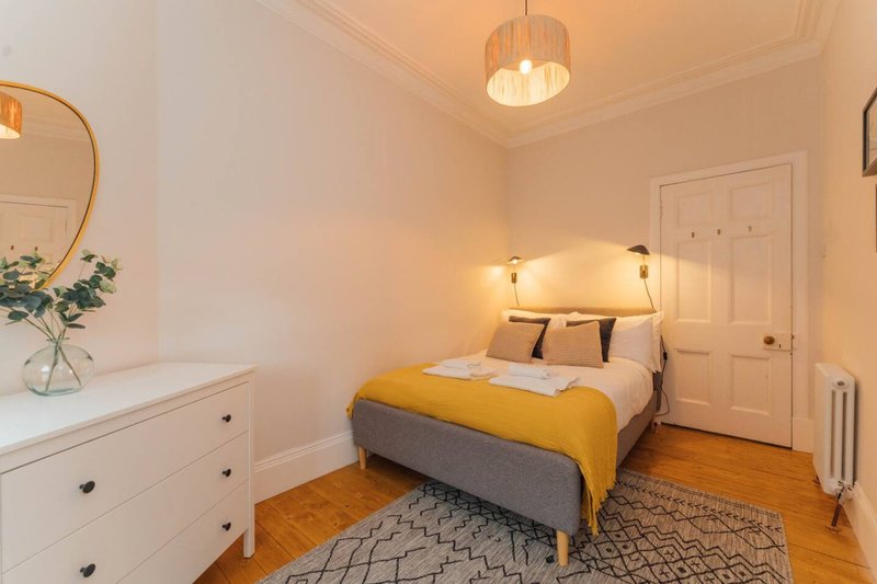 Bright bedroom with double bed, yellow throw, white dresser and round mirror