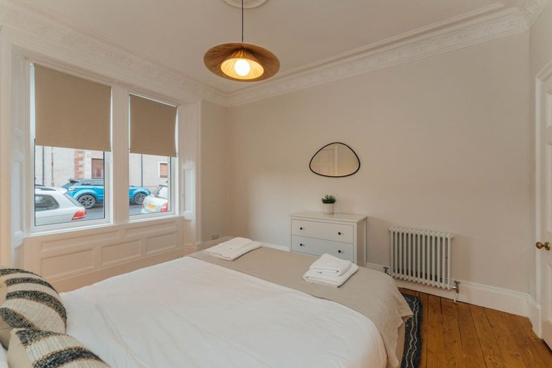 Bright bedroom with double bed, white dresser, radiator and large window overlooking street
