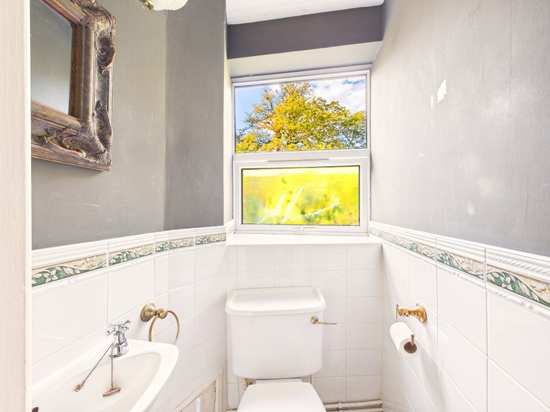 Small bathroom with white tiles, toilet, sink, and window overlooking garden