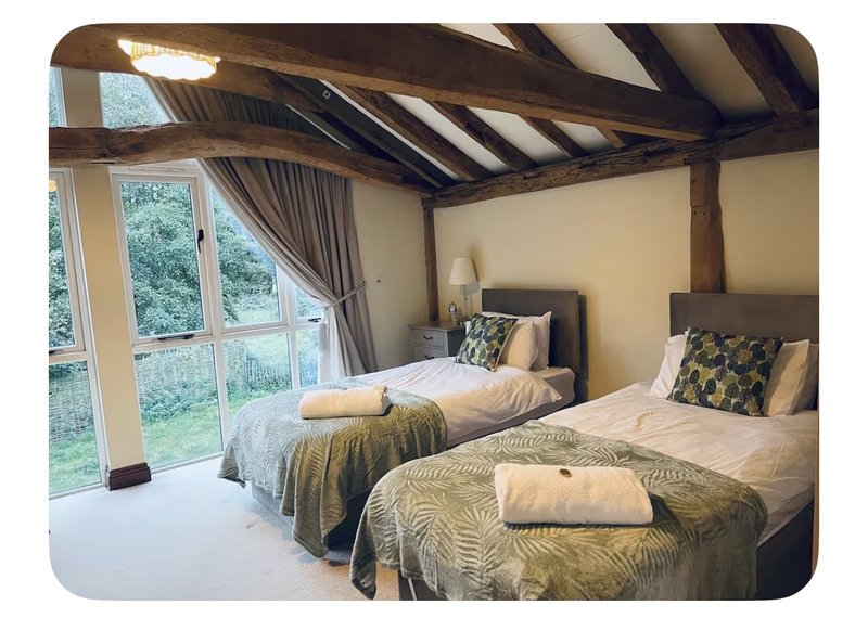 Spacious bedroom with exposed wooden beams, two beds with patterned bedding, and large windows overlooking garden