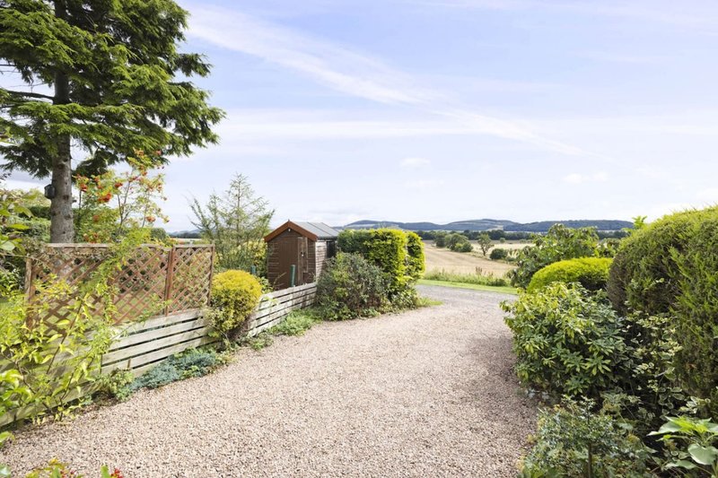 Gravel driveway with wooden gate, garden sheds, hedges and countryside hills beyond