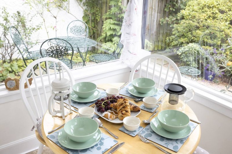 Bright dining area with set table, green dishes, pastries, and garden views through windows