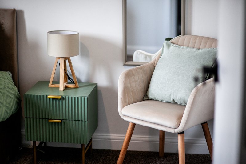 Modern bedroom corner with upholstered armchair, green bedside table, lamp, and cushions