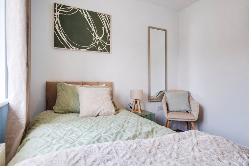 Bright bedroom with double bed, green patterned duvet, cushions, wall art, and wooden mirror