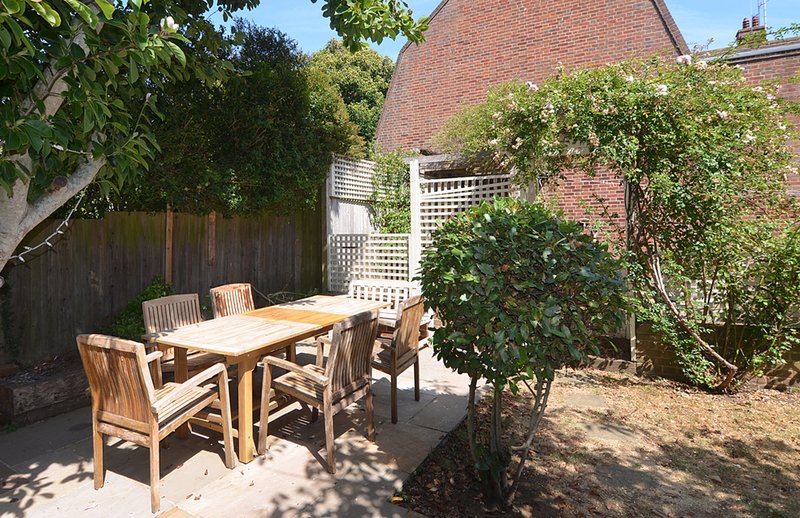 Garden patio with wooden dining table, chairs, ivy-covered trellis and brick house