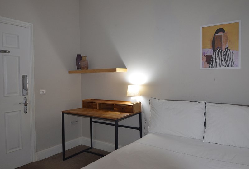 Bedroom with white bed, wooden desk with black frame, shelf, and framed artwork on wall