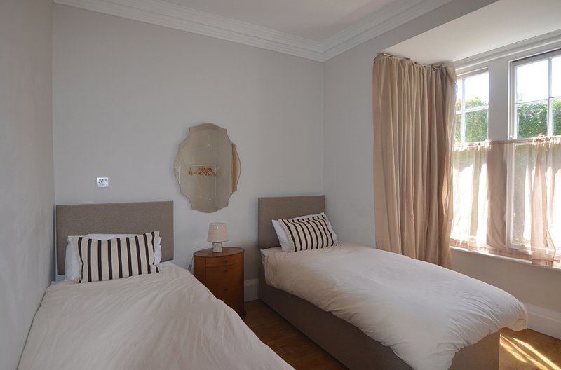 Twin bedroom with two single beds, wooden bedside table, round mirror and large windows with curtains