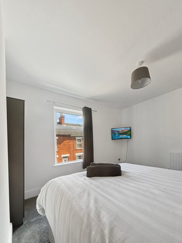 Bright bedroom with white bed, wall-mounted TV, pendant light and window overlooking brick building