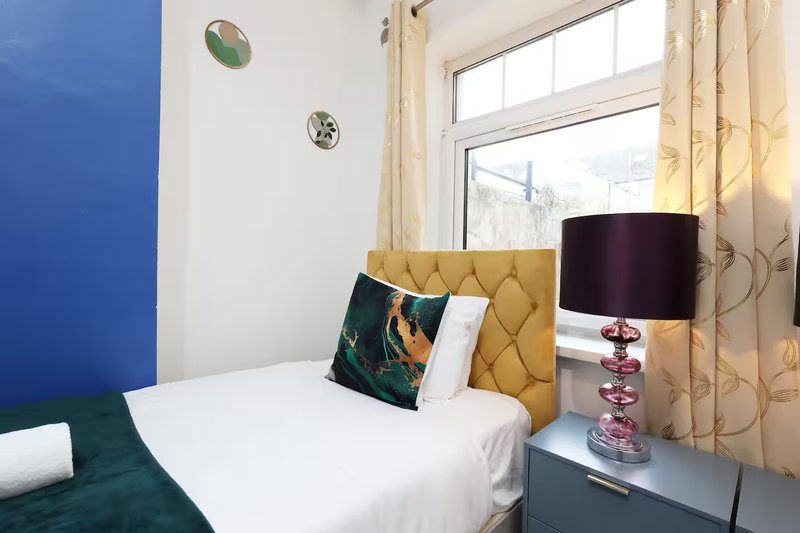 Bright bedroom with single bed, yellow headboard, bedside lamp and window with curtains